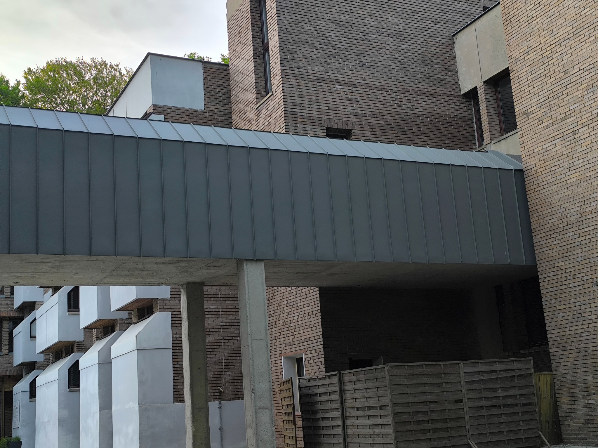 Szary, metalowy łącznik dachowy między budynkami z cegły, wsparty na betonowych kolumnach, z widocznymi elementami konstrukcyjnymi i fragmentem zieleni w tle.