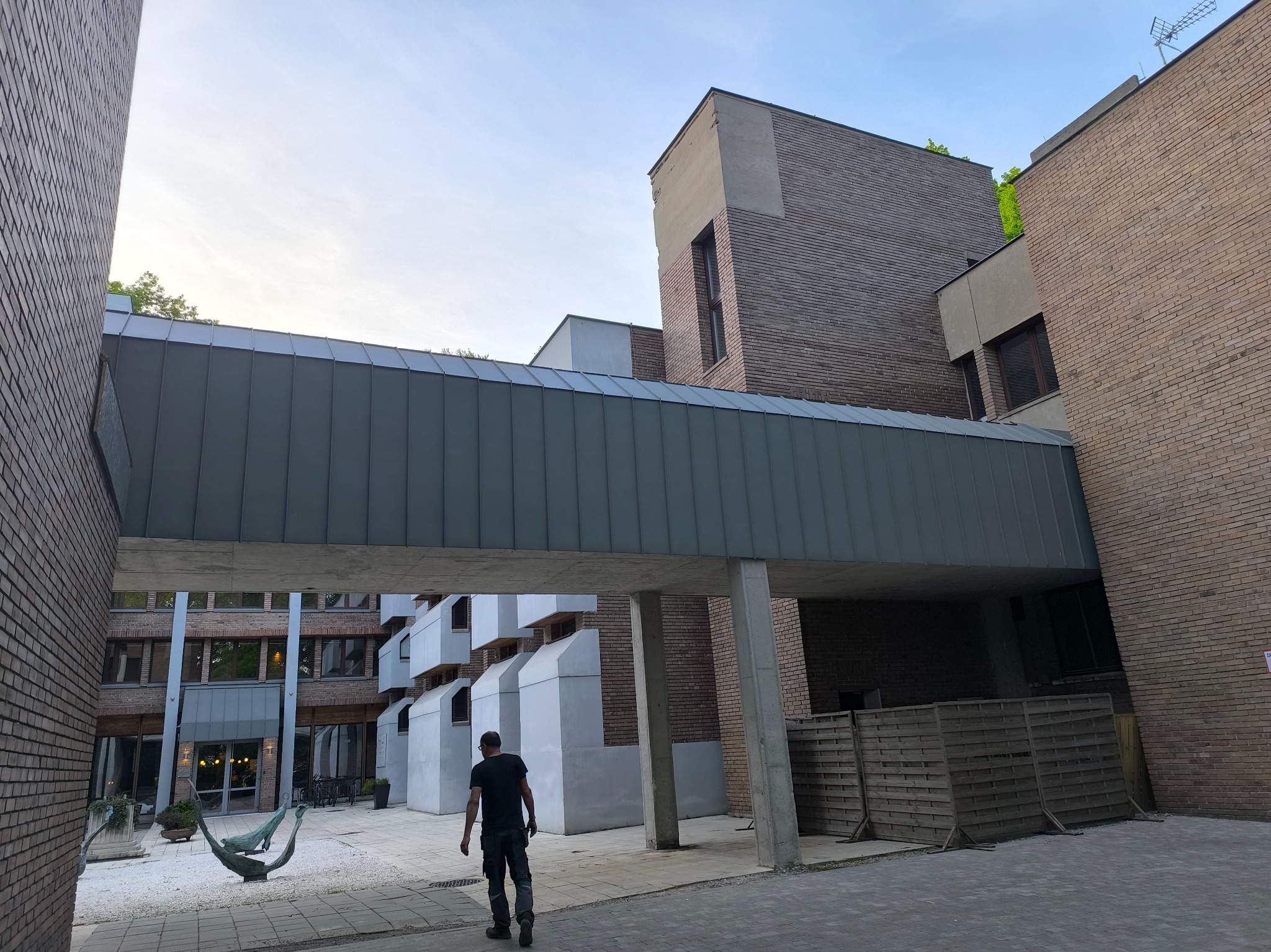 Szary, metalowy łącznik dachowy między budynkami z cegły, widziany z perspektywy pieszego. W tle dziedziniec z hamakiem.