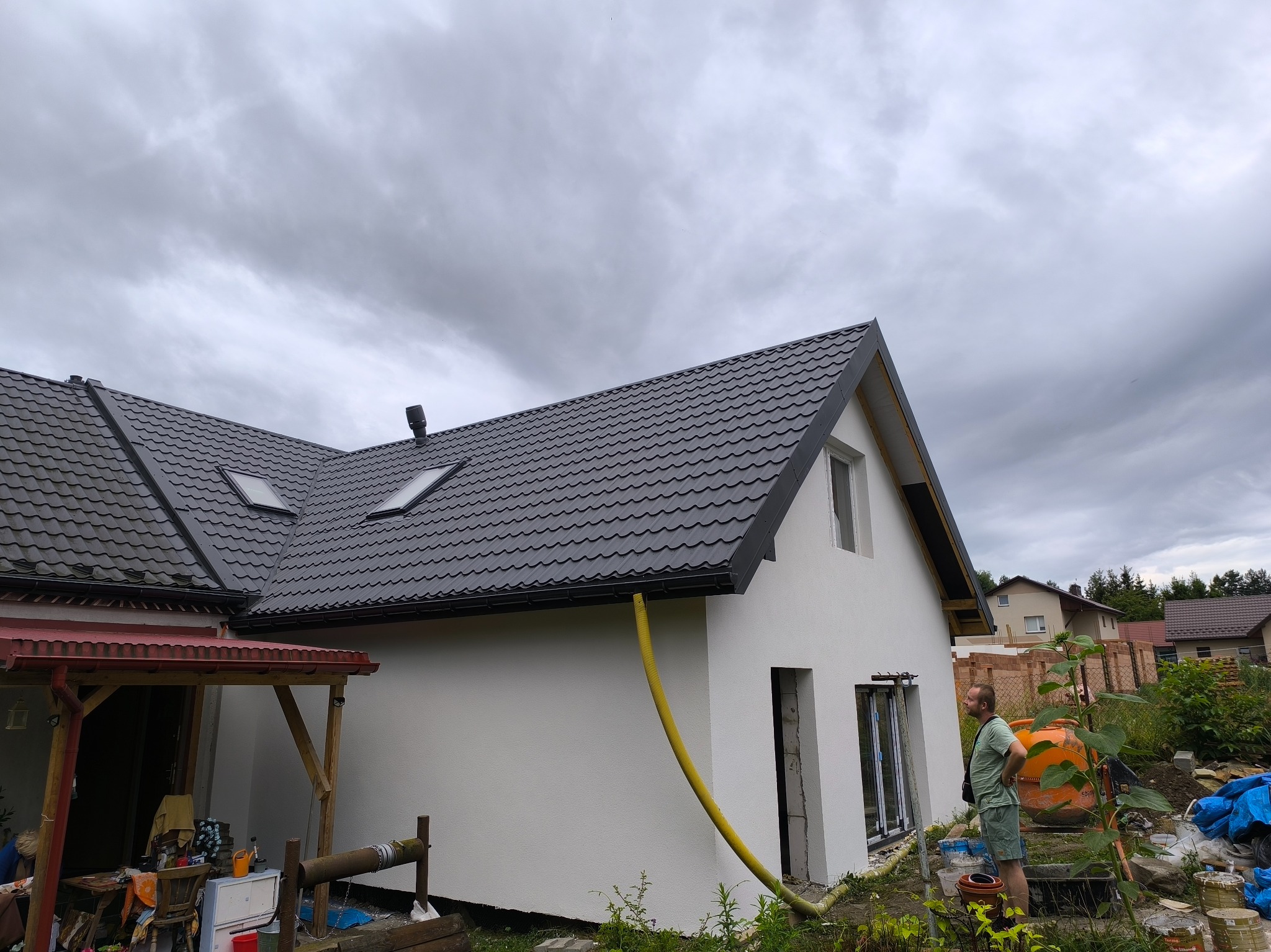 Dom z szarą dachówką i żółtą rurą spustową. Widoczny fragment elewacji, okna w trakcie montażu i osoba stojąca obok pomarańczowej betoniarki. Zachmurzone niebo w tle.