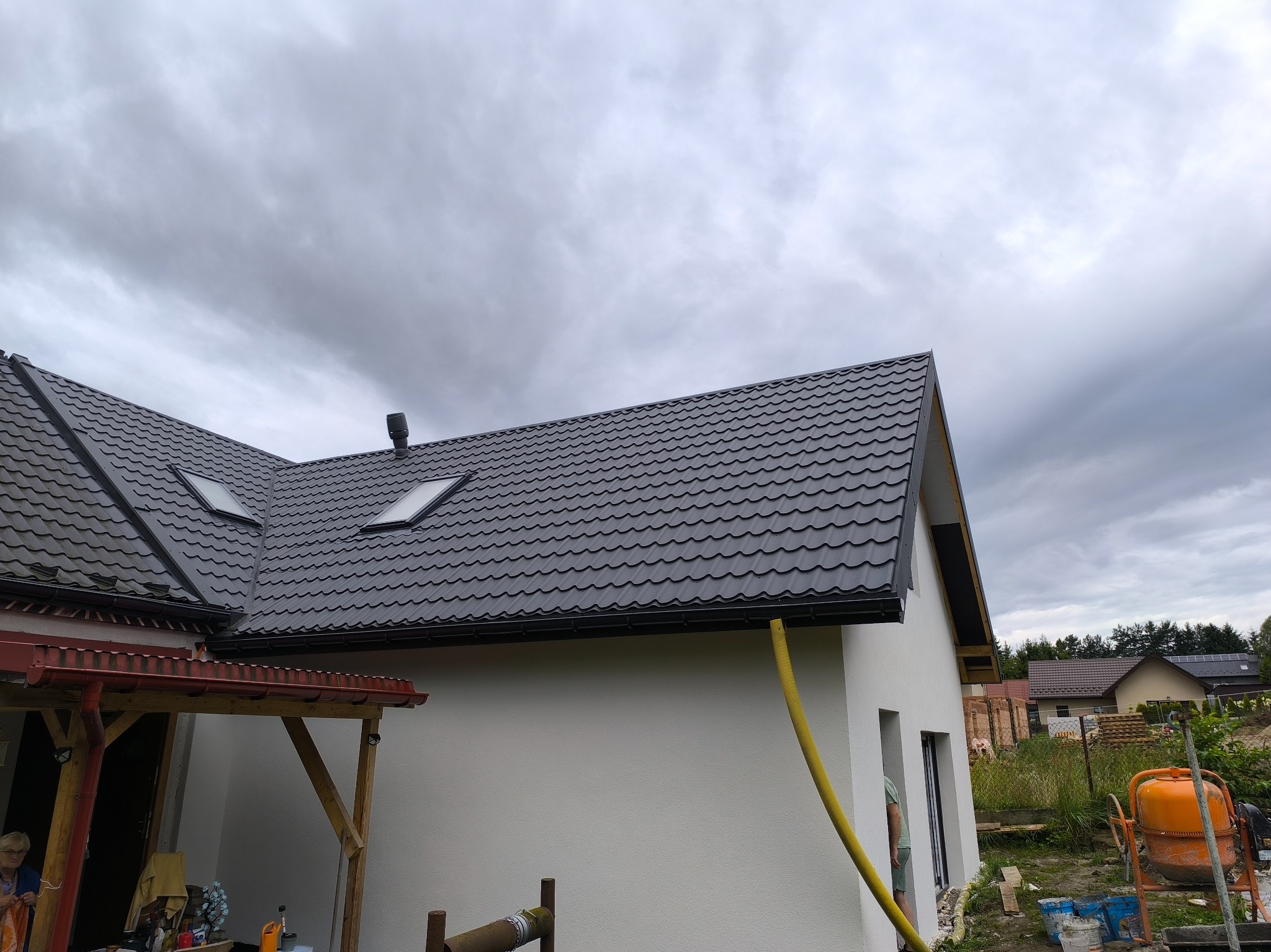 Nowy, grafitowy dach z oknami połaciowymi na białym domu jednorodzinnym. Widoczna rynna i żółty wąż. Na drugim planie inne domy i pochmurne niebo.