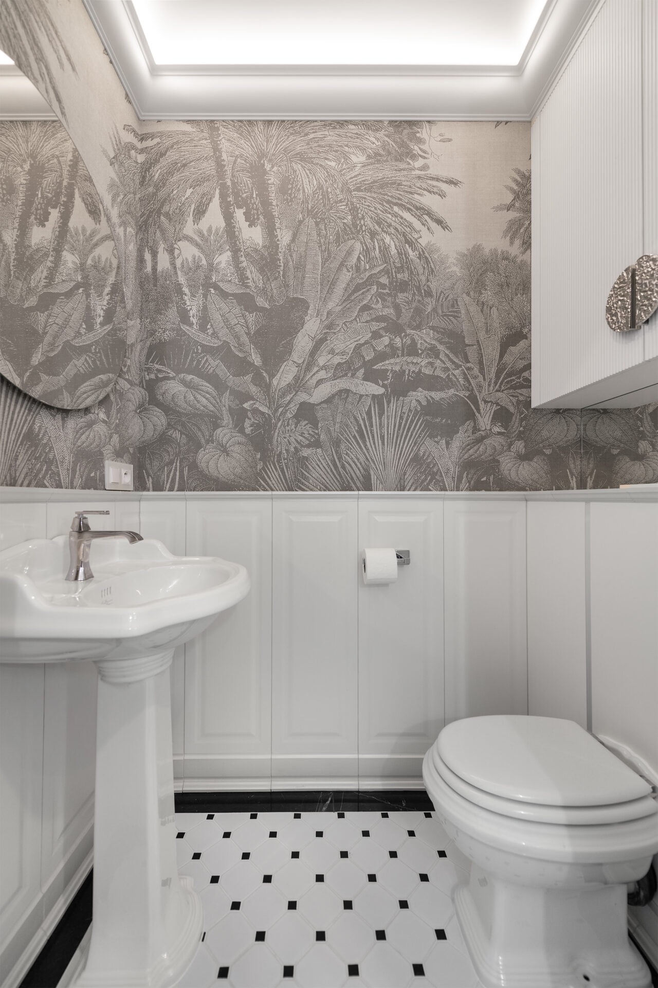Elegancka łazienka w stylu retro: biała ceramika, wzorzysta tapeta z motywem tropikalnym, czarno-biała podłoga. Umywalka na postumencie, toaleta i szafka.