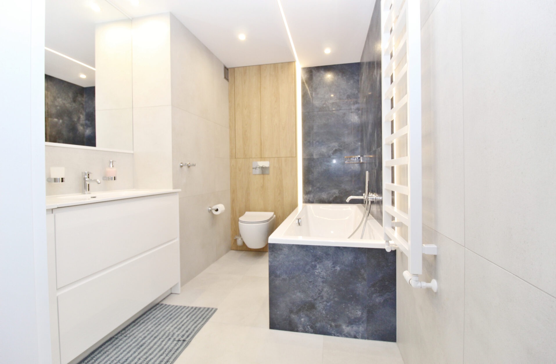 Nowoczesna łazienka z wanną obudowaną ciemnym gresem, białą umywalką z szafką i grzejnikiem drabinkowym. Jasne płytki i oświetlenie LED.
