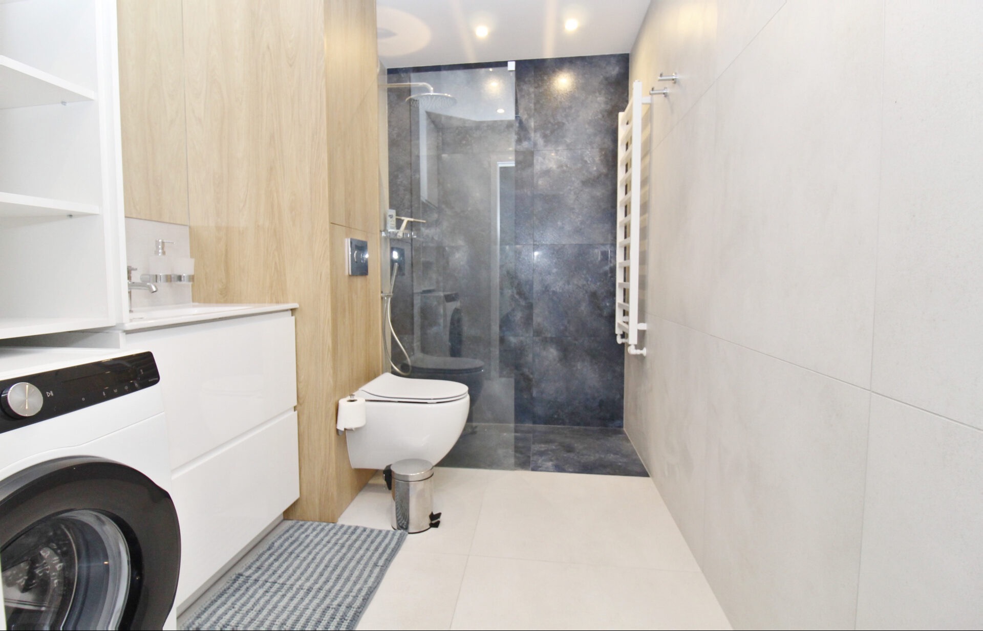 Nowoczesna łazienka z kabiną prysznicową walk-in, toaletą podwieszaną i pralką. Dominuje biel i drewno, akcentują szare płytki pod prysznicem.