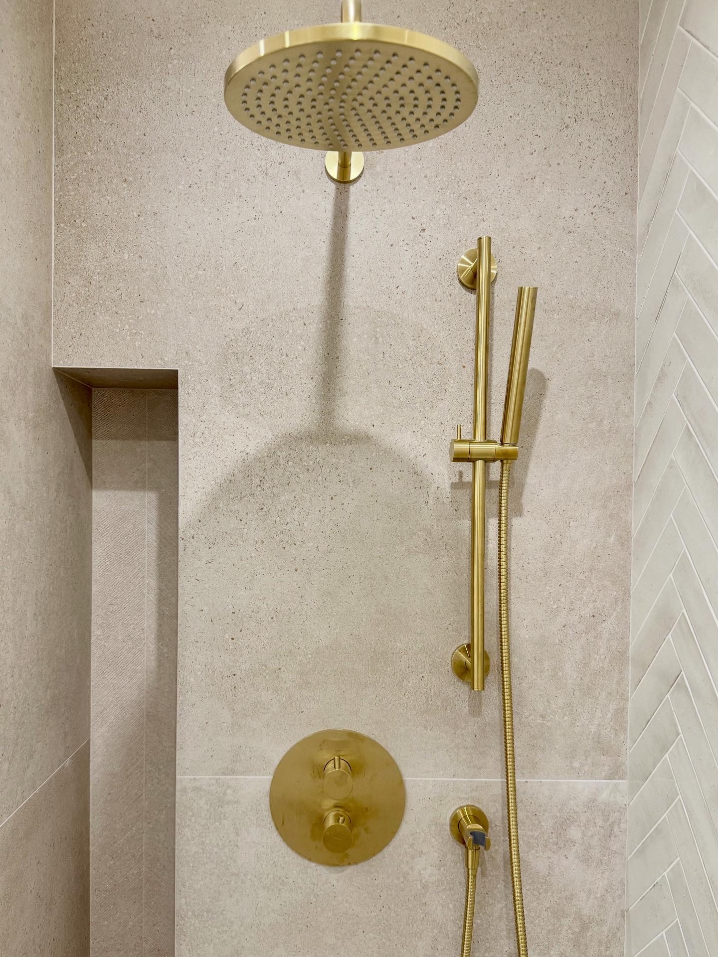 Beżowa ściana z złotą deszczownicą, baterią i słuchawką prysznicową. Płytki imitujące kamień i wzór jodełki. Nowoczesne wykończenie łazienki.