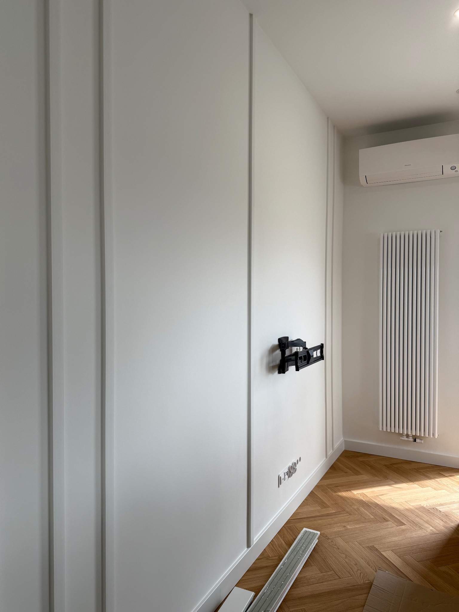 Białe ściany z dekoracyjnymi listwami, zamontowany uchwyt TV, grzejnik żeberkowy i parkiet w jodełkę. Widoczne gniazdka elektryczne i klimatyzator.