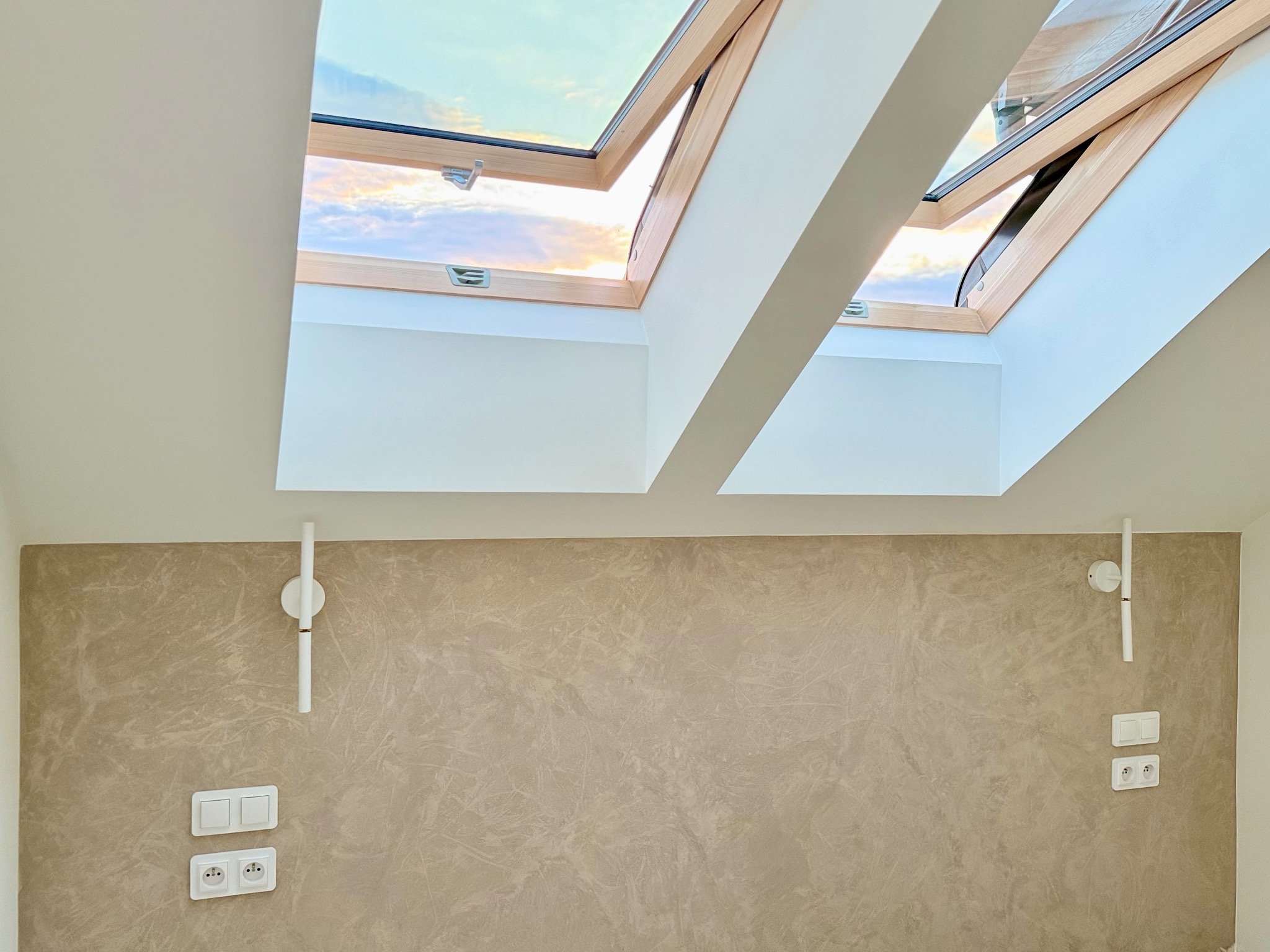 Wykończona ściana z efektem dekoracyjnym, lampy ścienne i okna dachowe z widokiem na niebo. Neutralna paleta barw, nowoczesny design.