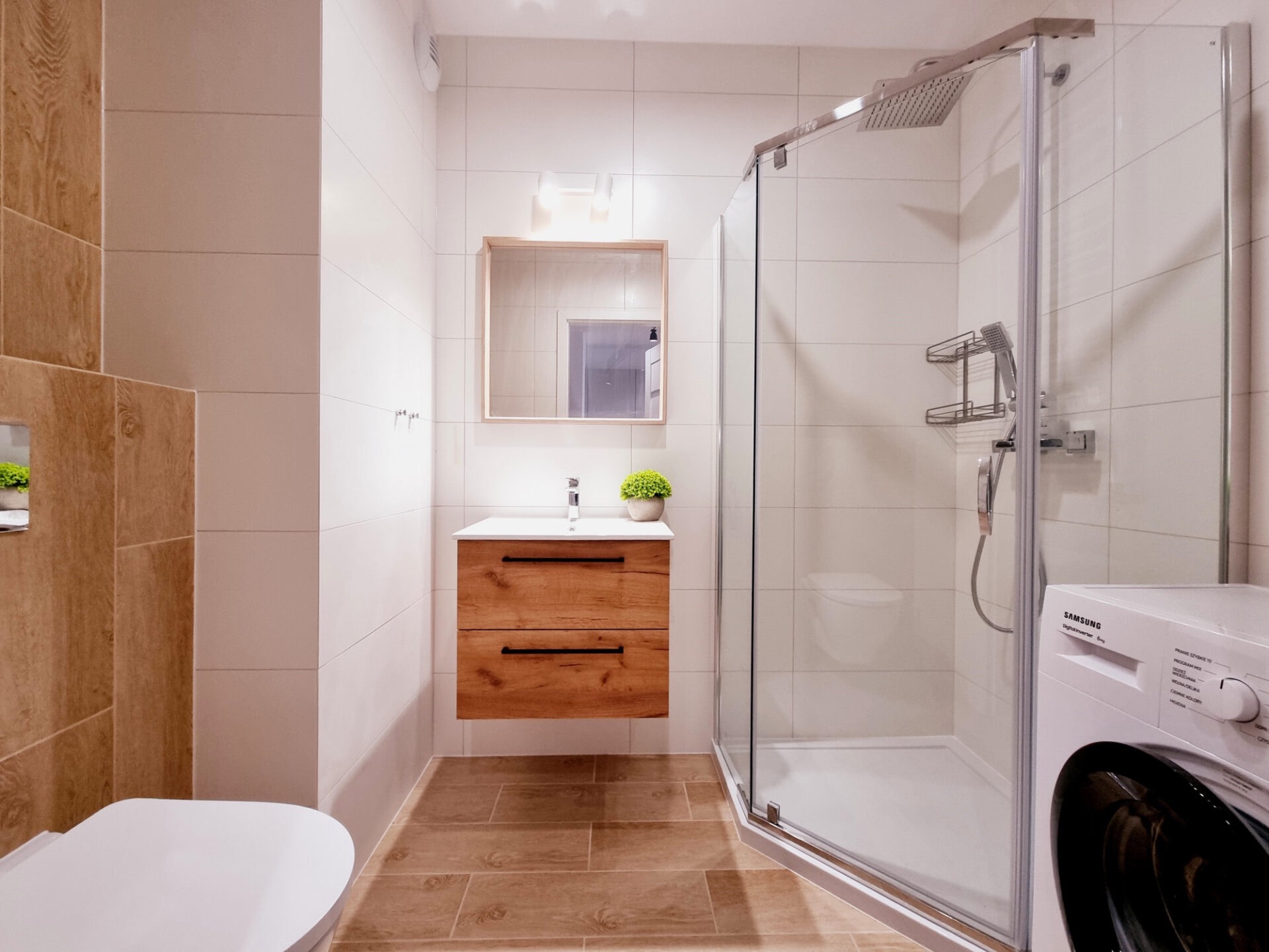 Nowoczesna łazienka z kabiną prysznicową, umywalką z szafką z drewna i pralką. Białe i drewniane płytki, lustro i roślina doniczkowa nadają wnętrzu przytulny charakter.