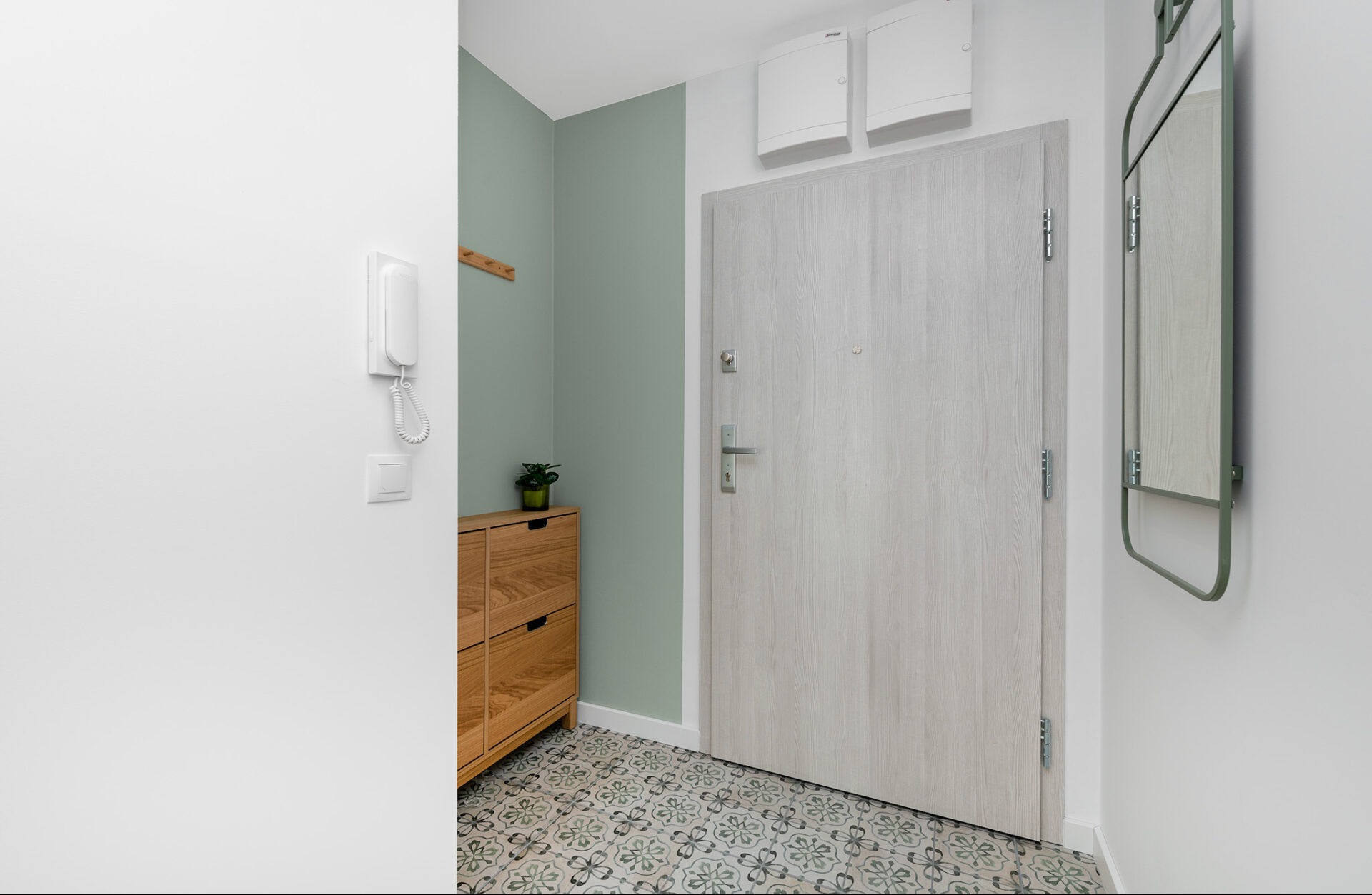 Jasne wnętrze przedpokoju z drewnianą szafką na buty, wzorzystą podłogą i szarymi drzwiami. Ściana w odcieniu zieleni. Nowoczesny design.