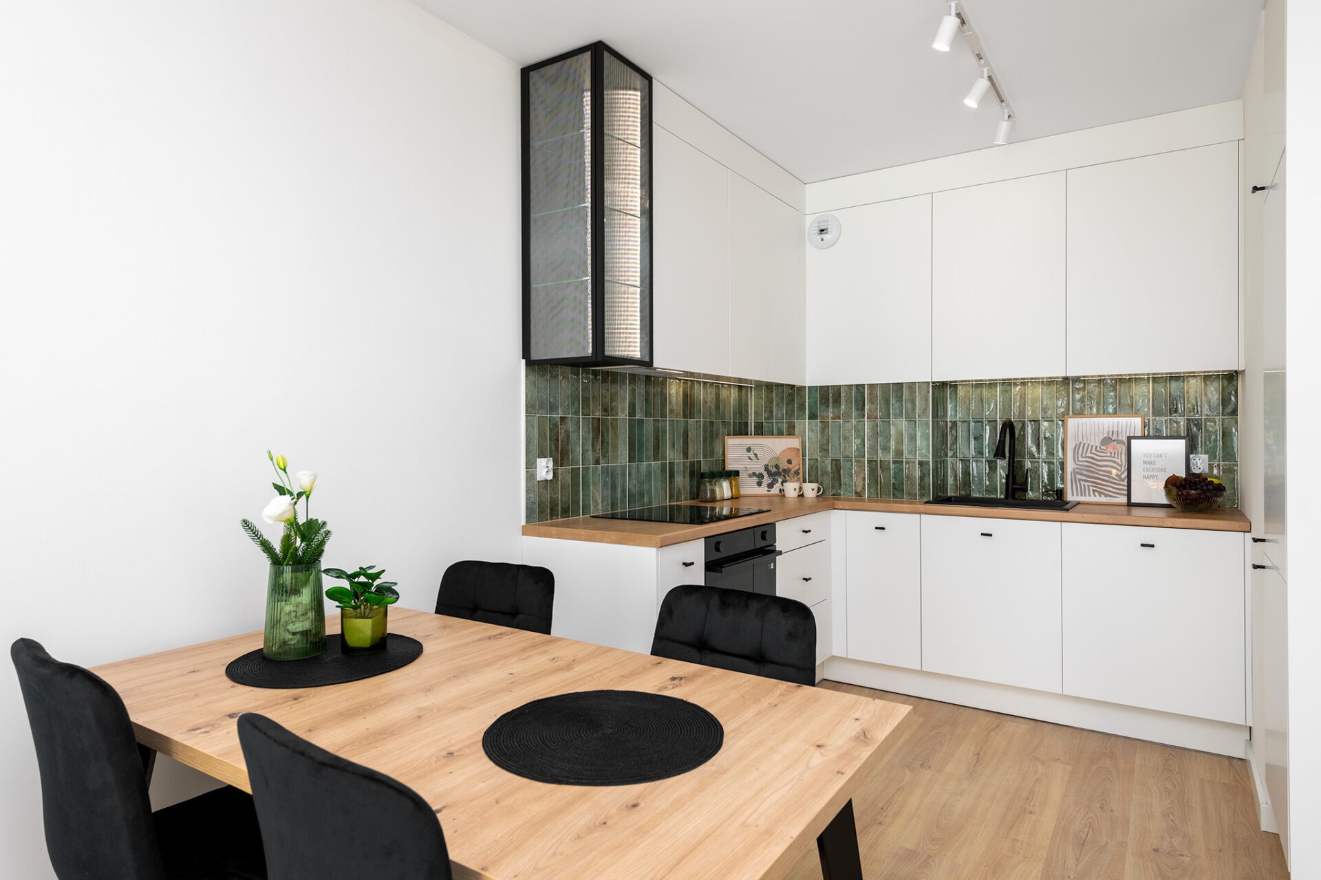 Nowoczesna kuchnia z zielonymi płytkami na ścianie, białymi szafkami i drewnianym blatem. Stół z czarnymi podkładkami i krzesłami. Oświetlenie punktowe.
