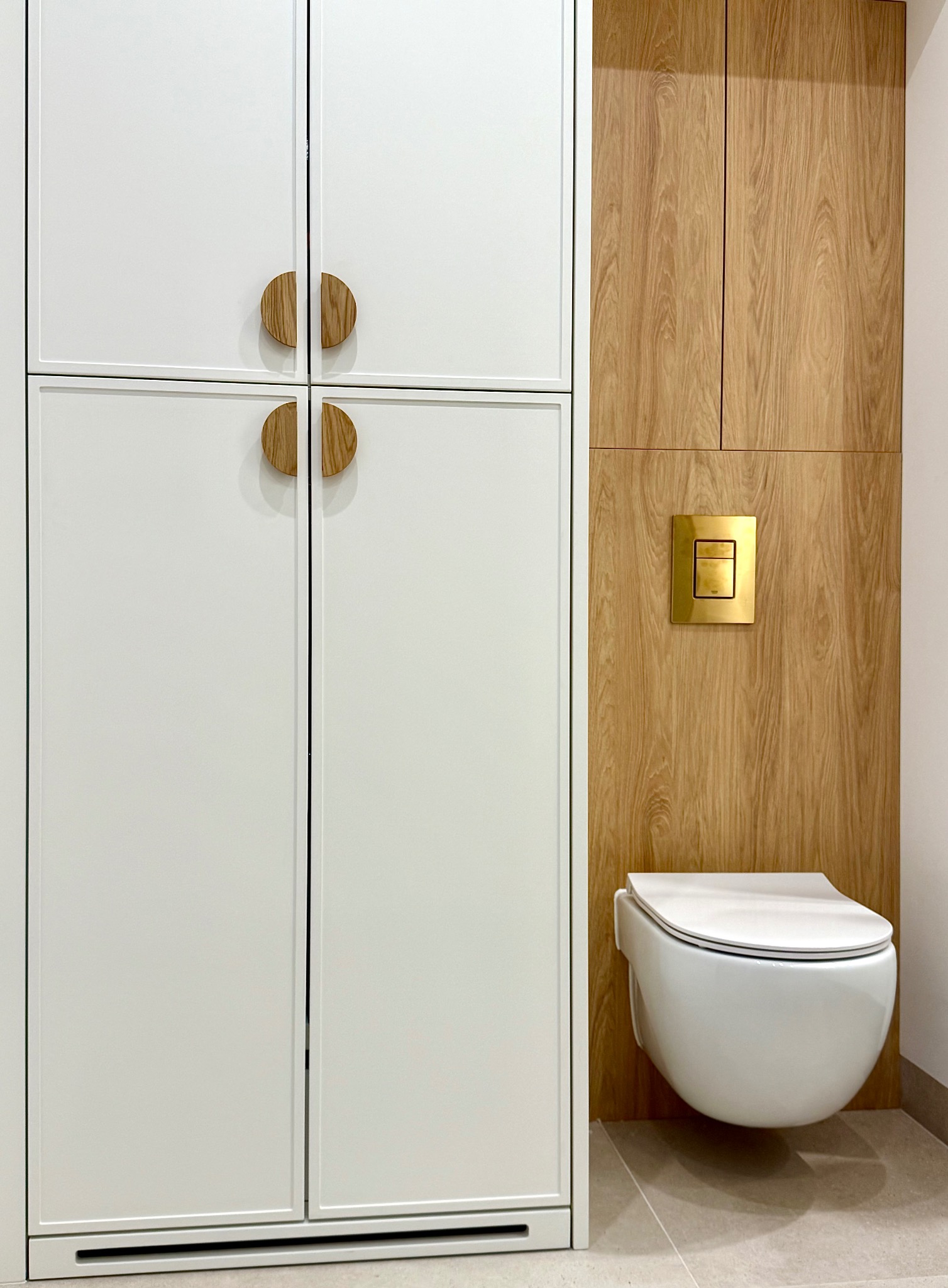 Nowoczesna toaleta podwieszana z drewnianą zabudową i złotym przyciskiem spłukującym, obok biała szafka z okrągłymi uchwytami. Minimalistyczny design.