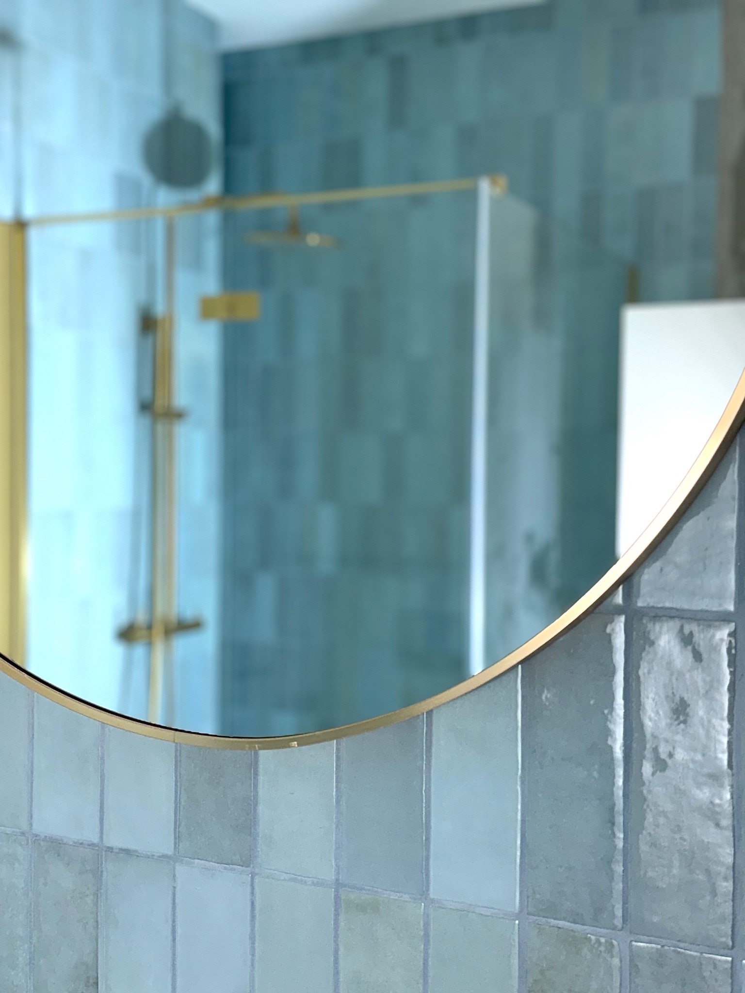 Błękitna łazienka z kabiną prysznicową i złotymi akcentami, widziana przez okrągłe lustro z ramą. Minimalistyczny design, płytki w odcieniach błękitu.