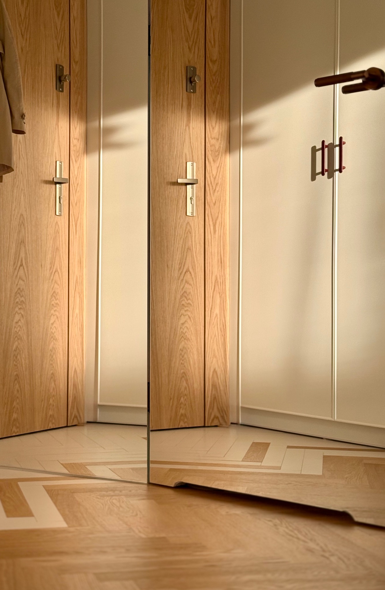 Nowoczesne wnętrze: drewniane drzwi z metalowymi klamkami, parkiet w geometryczny wzór, odbicie w lustrze. Ciepłe oświetlenie podkreśla detale wykończenia.