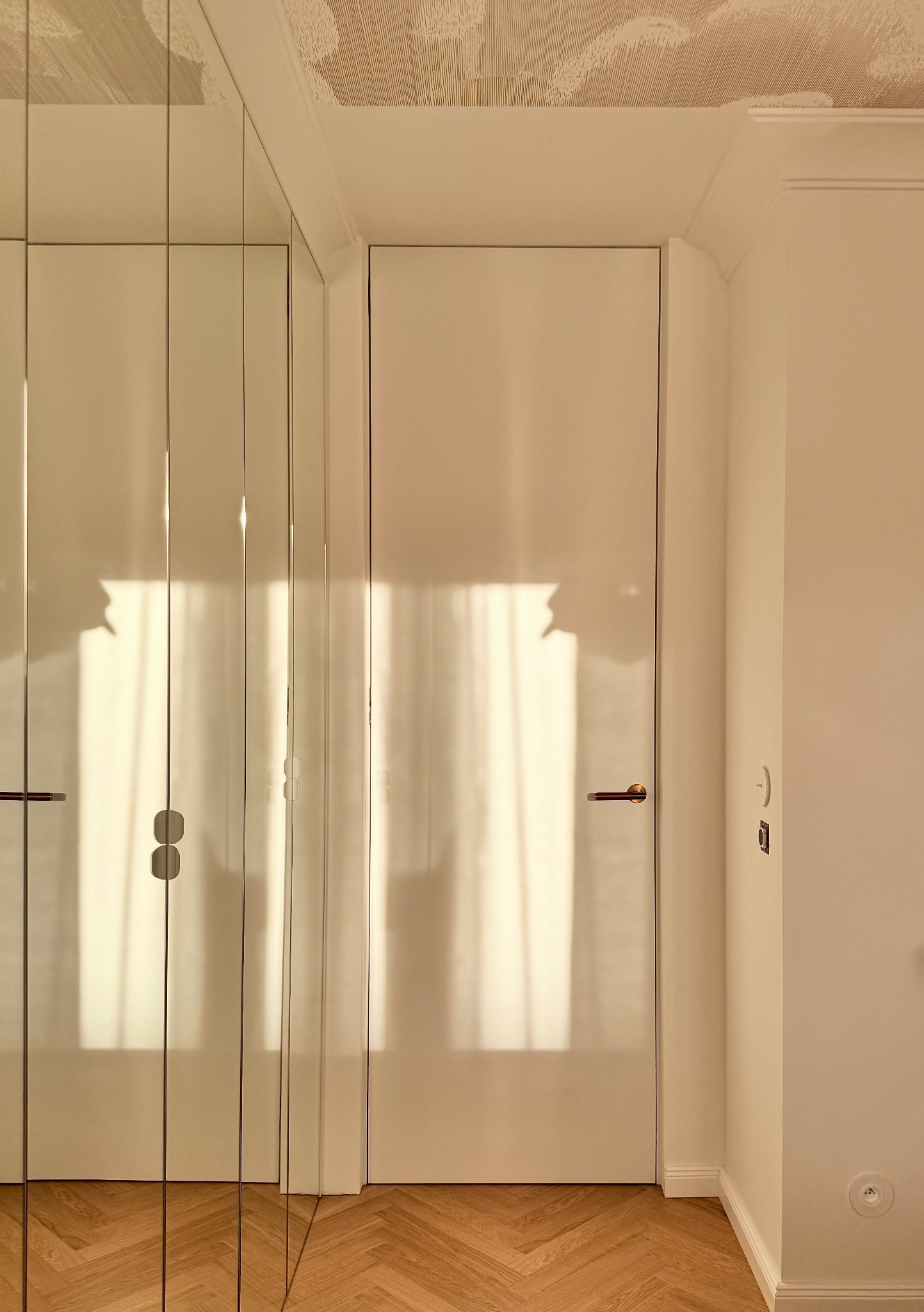 Białe drzwi wewnętrzne z mosiężną klamką, obok lustrzana szafa wnękowa, parkiet w jodełkę, kremowe ściany i sufit z delikatnym wzorem. Wnętrze po remoncie.