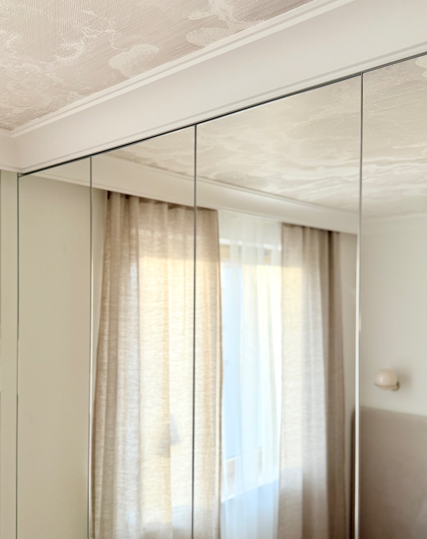 Wnętrze z lustrzanymi drzwiami szafy wnękowej, odbijającymi okno z zasłonami. Sufit z dekoracyjną fakturą. Minimalistyczny design.