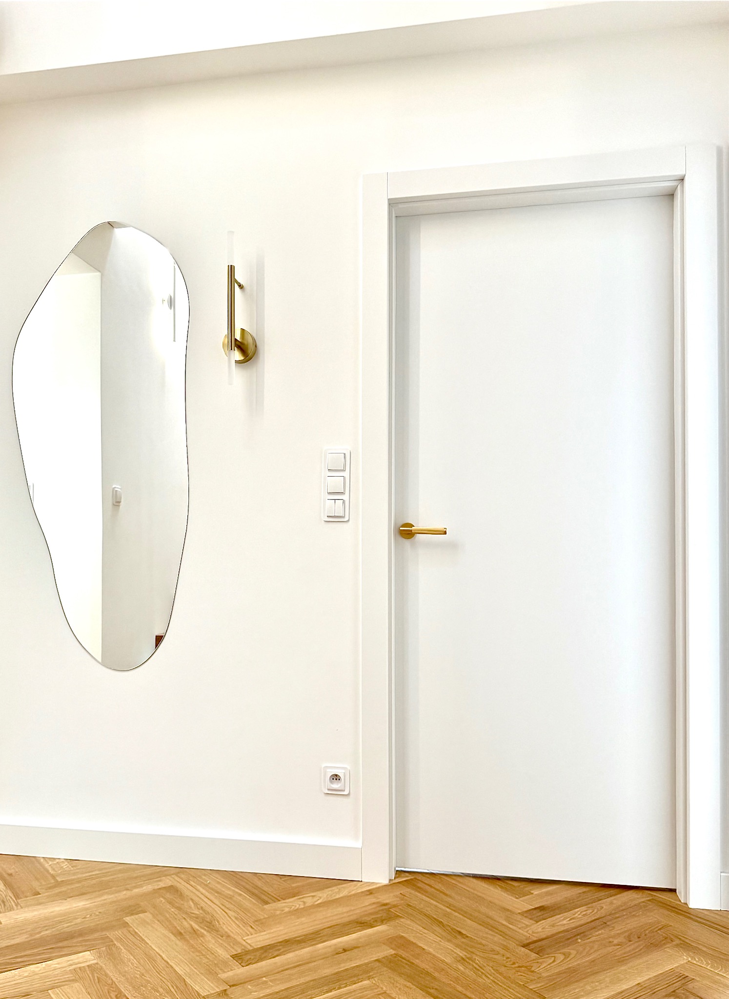 Białe drzwi z złotą klamką, asymetryczne lustro i lampa na ścianie w wykończonym wnętrzu z parkietem w jodełkę. Minimalistyczny design.