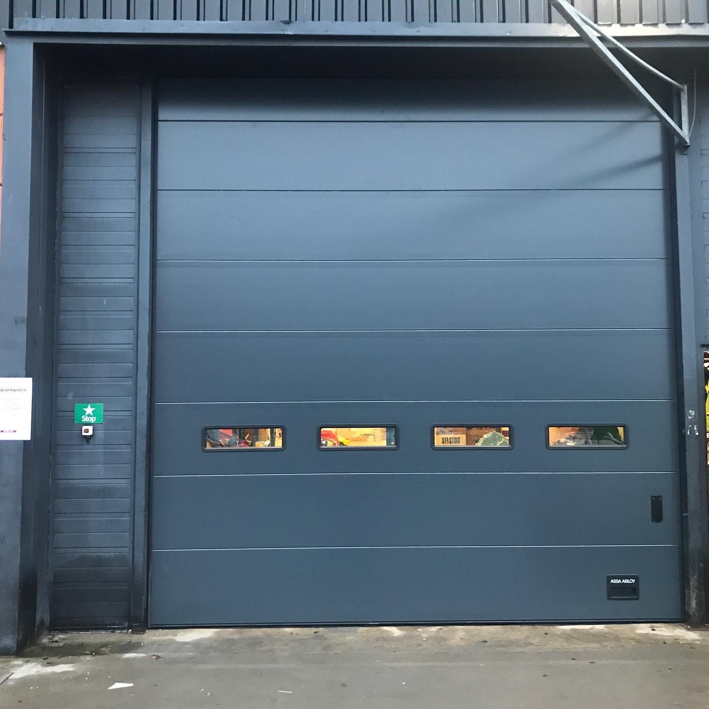 Szara brama segmentowa z trzema małymi oknami w hali przemysłowej. Widoczne logo ASSA ABLOY w prawym dolnym rogu. Na lewo od bramy znak Stop z gwiazdą.