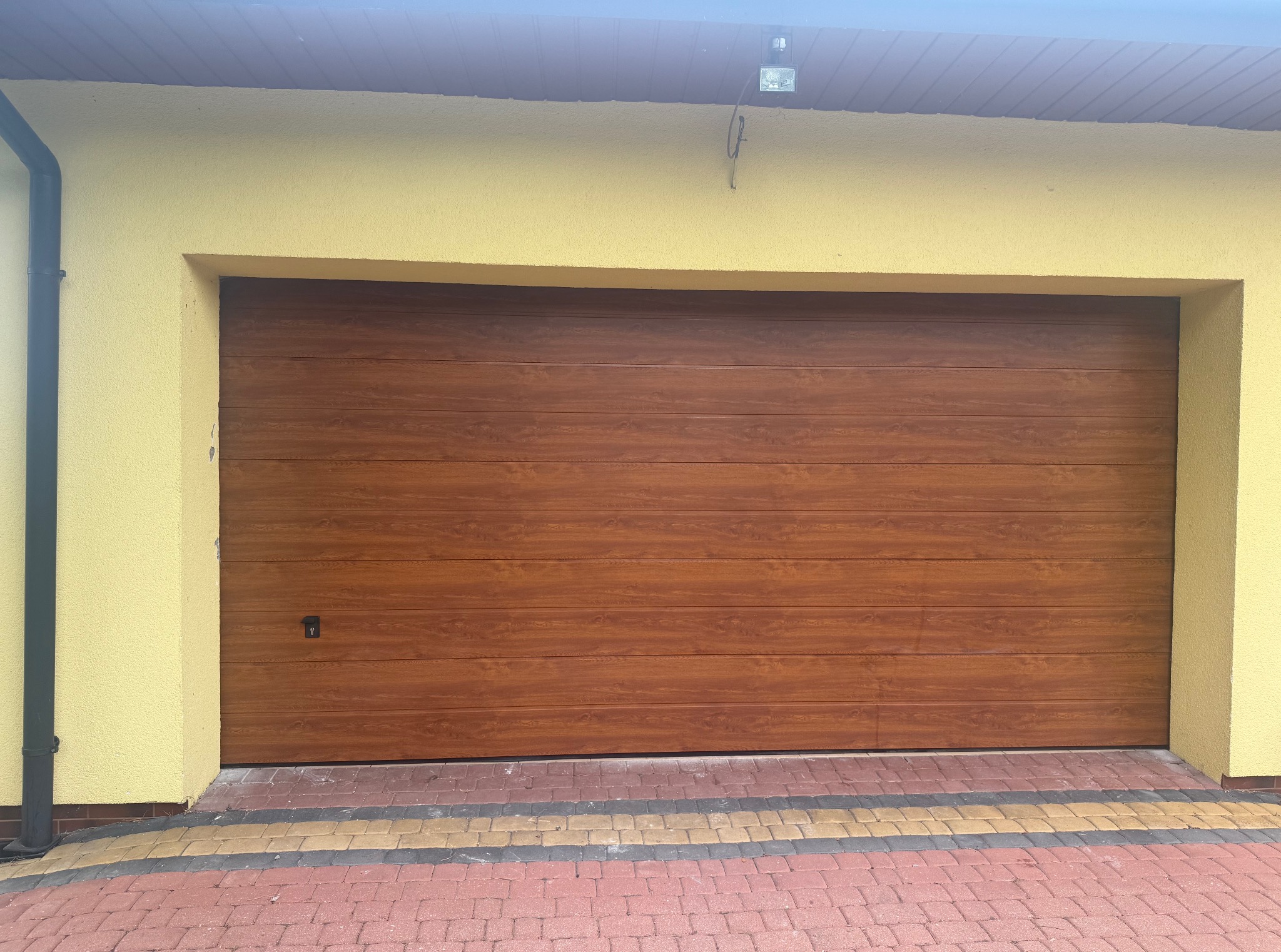 Zewnętrzny widok na zamkniętą, segmentową bramę garażową z imitacją drewna, zamontowaną w żółtym budynku z kostką brukową w trzech kolorach.