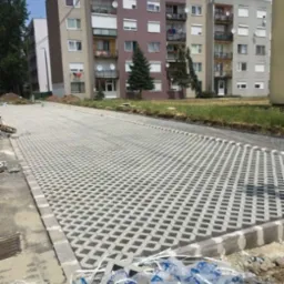 Układanie ażurowej kostki brukowej na podjeździe przed blokiem mieszkalnym. Widoczne resztki folii po kostce. Praca w toku, częściowo ułożona nawierzchnia.