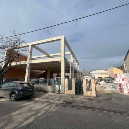 Road-Ép General Kft - Surowy szkielet betonowy dwukondygnacyjnego domu w trakcie budowy, widok z ulicy, z materiałami budowlanymi w tle i zaparkowanym samochodem.