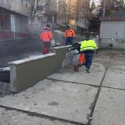Road-Ép General Kft - Ekipa w jaskrawych strojach tynkuje betonowy murek z kamienną górą. Częściowo otynkowana ściana, betonowy plac i budynki w tle. Prace wykończeniowe na zewnątrz.
