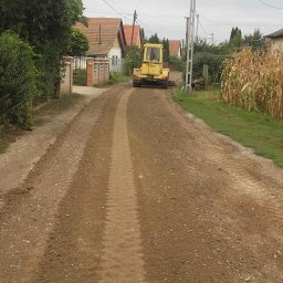 Road-Ép General Kft - Żółty walec drogowy wyrównuje gruntową drogę w wiejskiej scenerii. Widoczne domy z czerwonymi dachami i słup elektryczny. Ślady opon na ziemi.