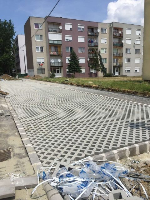 Układanie ażurowej kostki brukowej na podjeździe przed blokiem mieszkalnym. Widoczne resztki folii po kostce. Praca w toku, częściowo ułożona nawierzchnia.