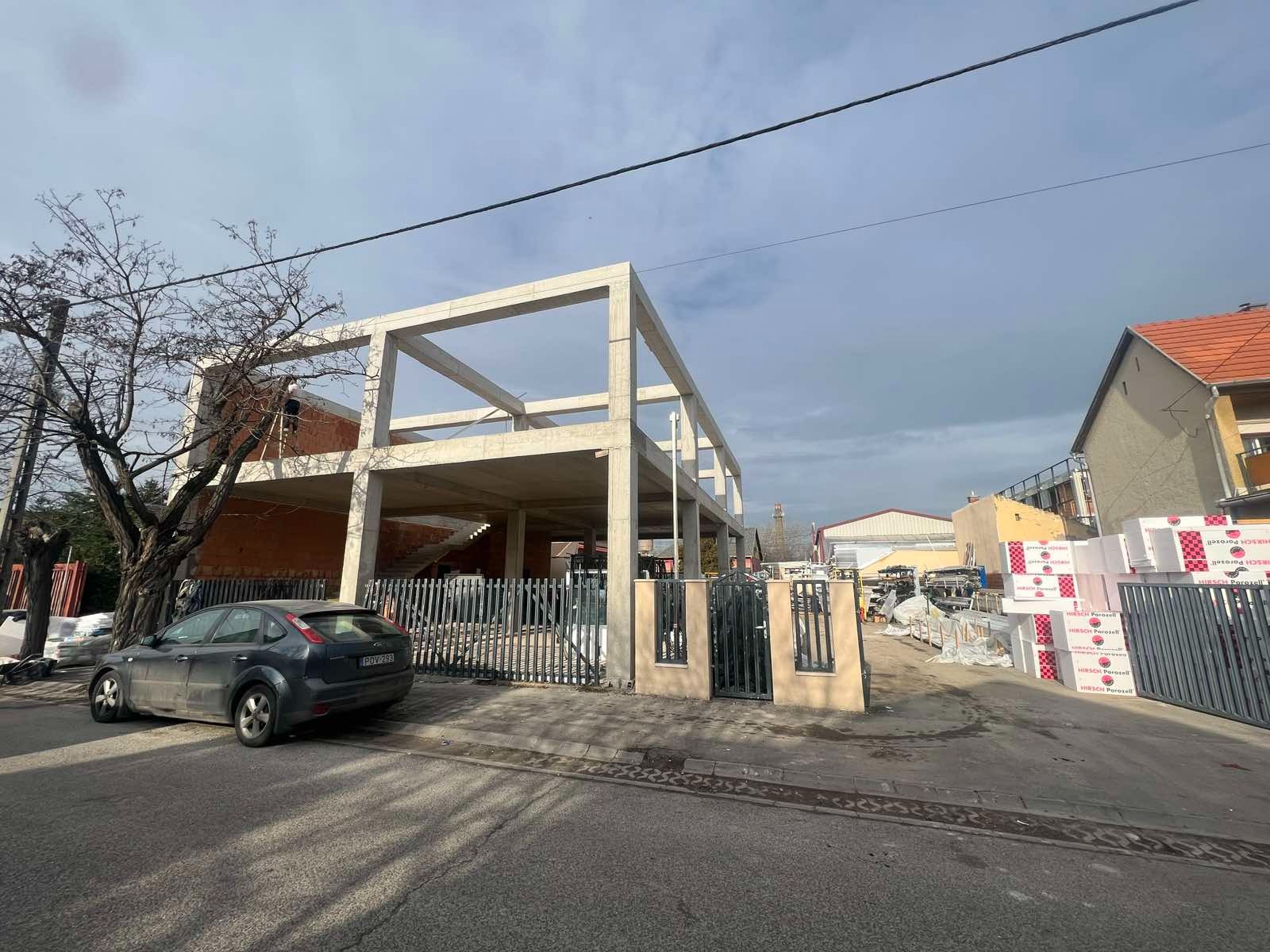 Surowy szkielet betonowy dwukondygnacyjnego domu w trakcie budowy, widok z ulicy, z materiałami budowlanymi w tle i zaparkowanym samochodem.