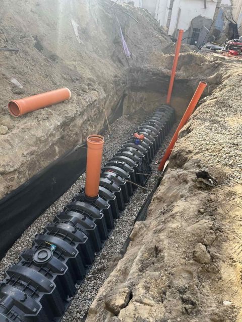 Układanie drenażu rozsączającego w wykopie. Widoczne czarne moduły drenażowe, rury pomarańczowe i obsypka żwirowa. Wykop zabezpieczony geowłókniną.