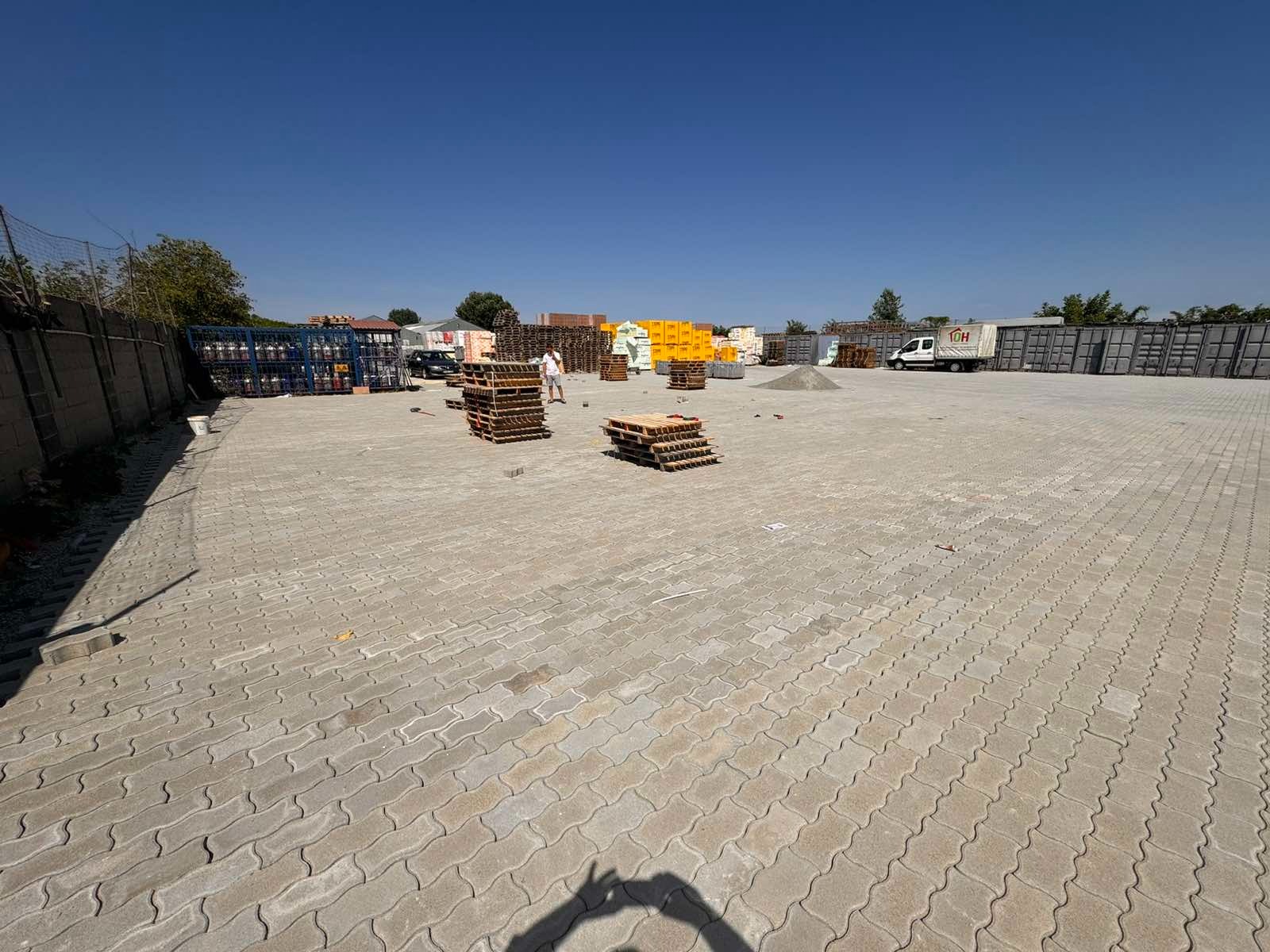 Plac wyłożony szarą kostką brukową typu fala, z paletami materiałów budowlanych w tle i wyraźnym cieniem osoby robiącej zdjęcie w dolnej części kadru.