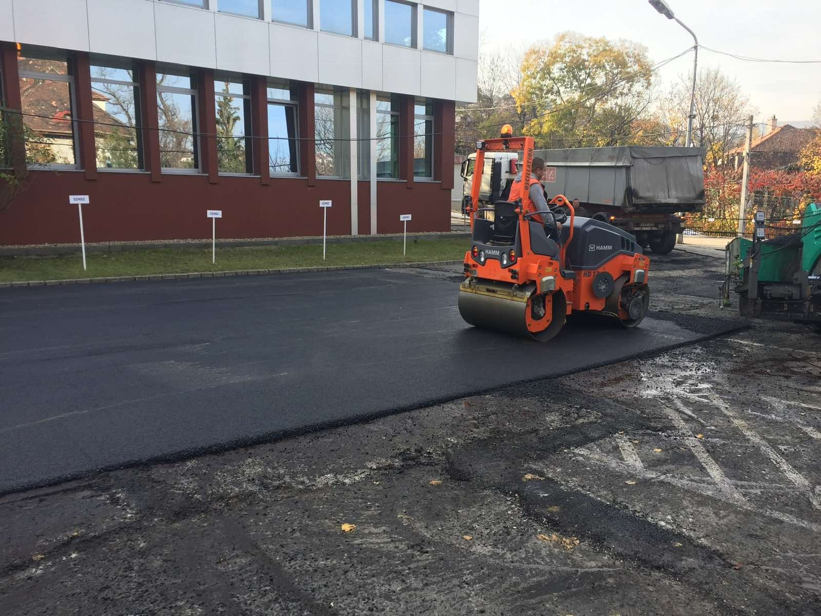 Walec drogowy HAMM HD 12 podczas prac asfaltowych na parkingu, z widocznym budynkiem w tle i świeżo ułożoną nawierzchnią.