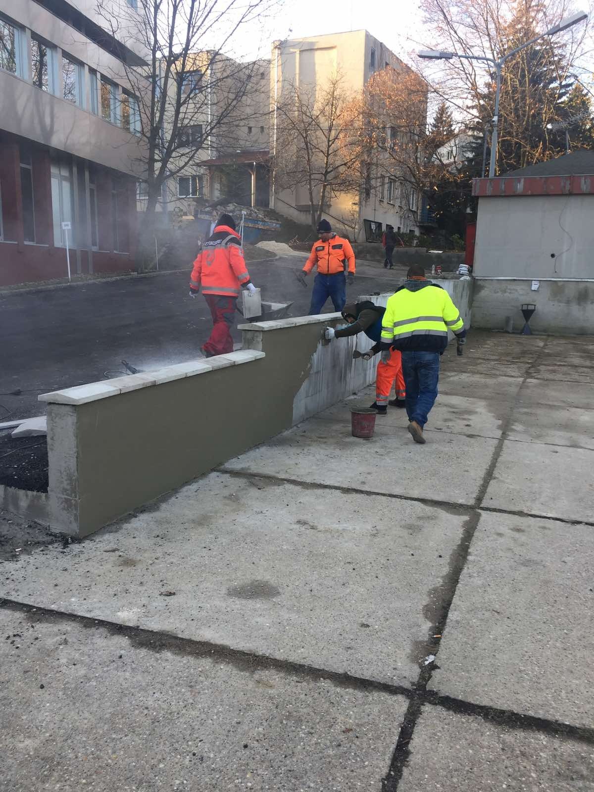 Ekipa w jaskrawych strojach tynkuje betonowy murek z kamienną górą. Częściowo otynkowana ściana, betonowy plac i budynki w tle. Prace wykończeniowe na zewnątrz.