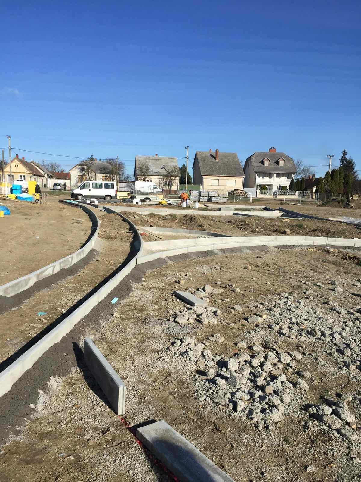 Fundamenty pod budowę chodnika na osiedlu domów jednorodzinnych, widok na ułożone krawężniki i przygotowany teren pod wylewkę, w tle budynki mieszkalne i błękitne niebo.