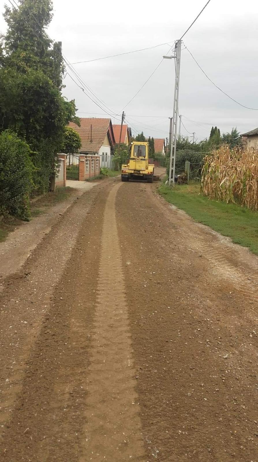 Żółty walec drogowy wyrównuje gruntową drogę w wiejskiej scenerii. Widoczne domy z czerwonymi dachami i słup elektryczny. Ślady opon na ziemi.