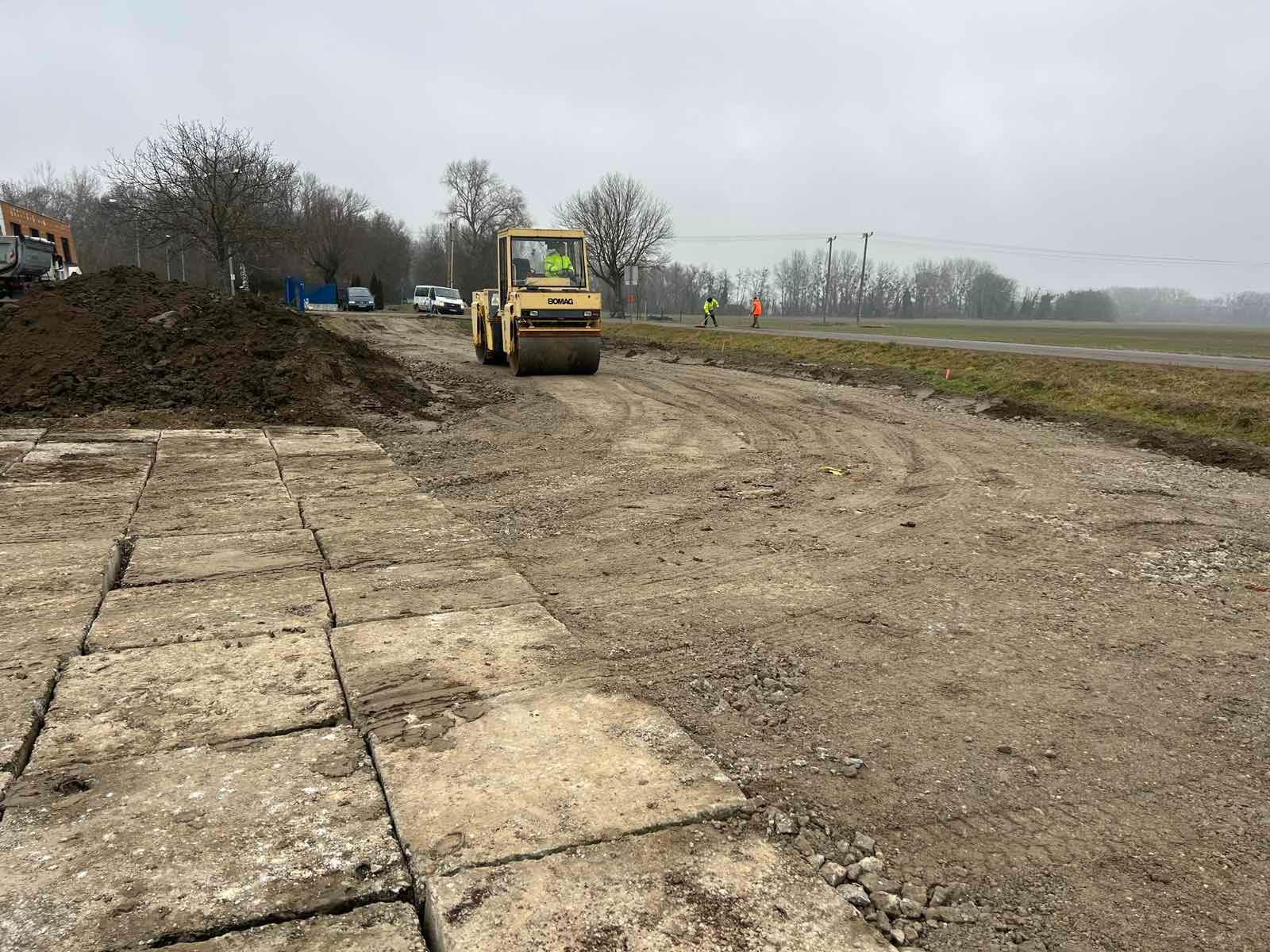 Żółty walec BOMAG ubija grunt na placu budowy, widoczne płyty betonowe, hałda ziemi i pracownicy w odblaskowych ubraniach. Pochmurny dzień.