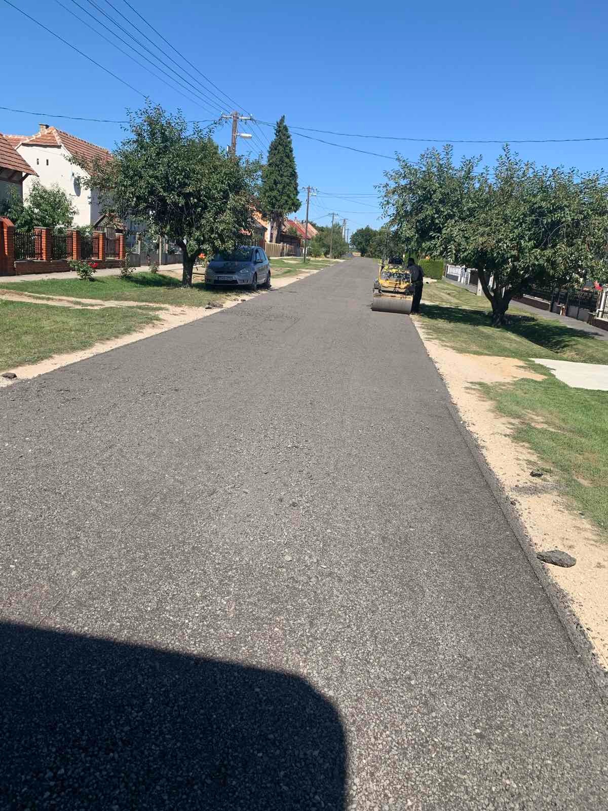 Świeżo wyasfaltowana ulica w obszarze zabudowanym, z walcem drogowym i pracownikiem w tle, drzewa po obu stronach drogi, błękitne niebo w słoneczny dzień.