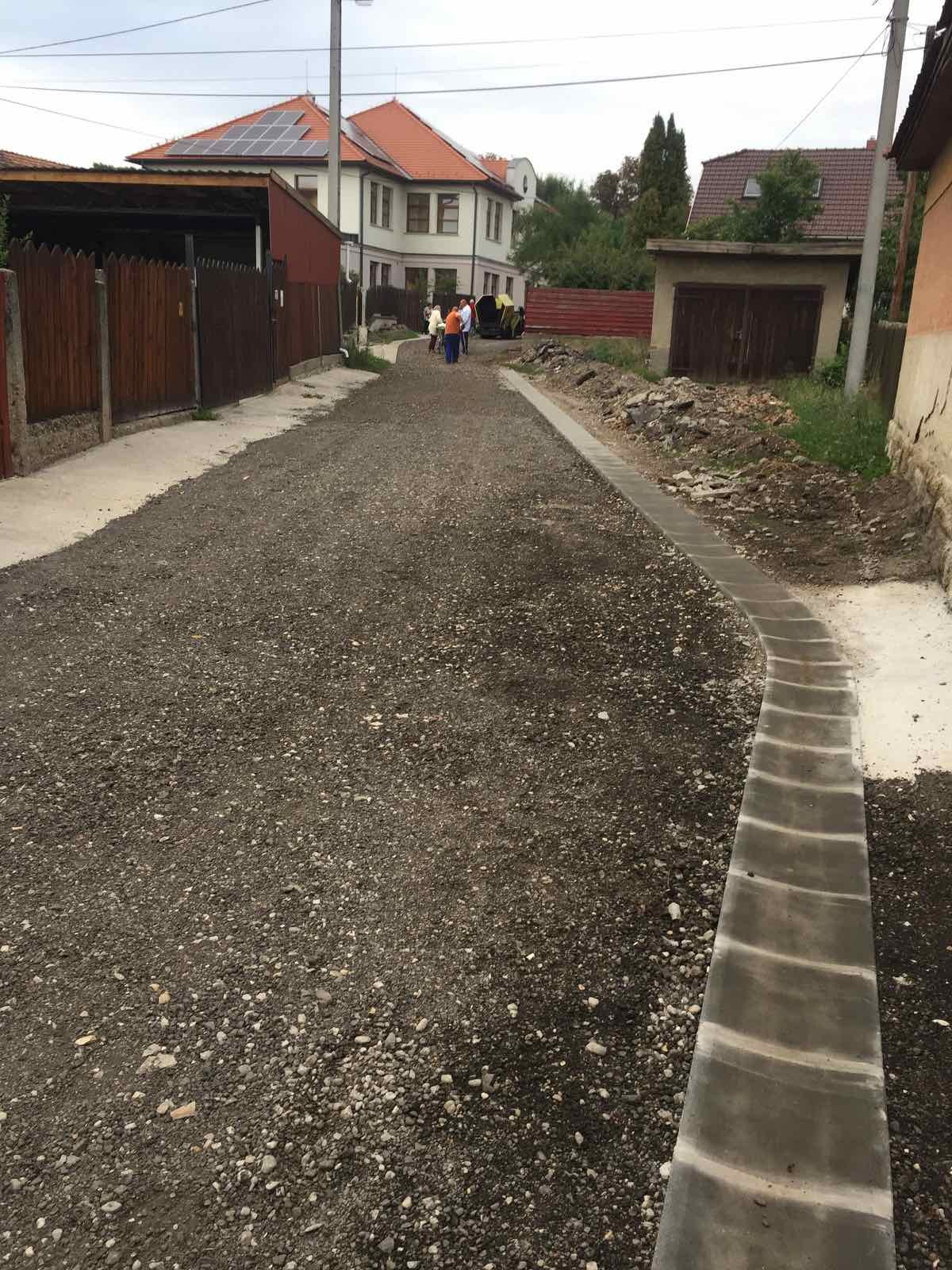 Ulica w trakcie budowy z nowym korytem odwadniającym. W tle domy, w tym jeden z panelami słonecznymi na dachu. Widoczne osoby i maszyna budowlana.