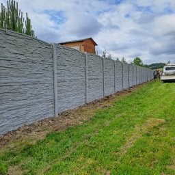 Markowe Ogrodzenia Betonowe Marek Januszewski - Długi, szary, betonowy płot z teksturą kamienia oddziela zielony trawnik od dalszej posesji. Widoczny fragment nieba z chmurami i budynek w tle.