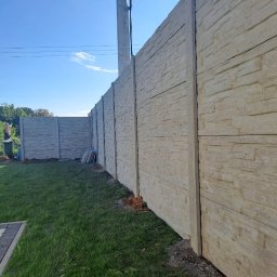 Markowe Ogrodzenia Betonowe Marek Januszewski - Długi, betonowy płot z fakturą kamienia, biegnący wzdłuż trawnika pod błękitnym niebem. Słup elektryczny w tle.