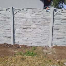 Markowe Ogrodzenia Betonowe Marek Januszewski - Eleganckie ogrodzenie betonowe w odcieniach szarości z dekoracyjnymi słupkami, otoczone świeżo obsypaną ziemią i trawą, z fragmentem dachu budynku w tle.