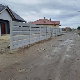 Markowe Ogrodzenia Betonowe Marek Januszewski - Długi, szary, betonowy płot panelowy oddzielający posesję od błotnistej drogi. Widoczne domy w tle i pochmurne niebo.