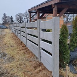 Markowe Ogrodzenia Betonowe Marek Januszewski - Nowoczesne ogrodzenie betonowe z poziomymi szczelinami, świeżo zamontowane, z widoczną ziemią i zielonymi tujami w tle, pod pochmurnym niebem.