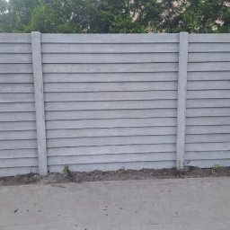 Markowe Ogrodzenia Betonowe Marek Januszewski - Szary, poziomy panel ogrodzeniowy z betonowymi słupkami, imitujący drewno, z zielonymi drzewami w tle i szarą kostką brukową u dołu.