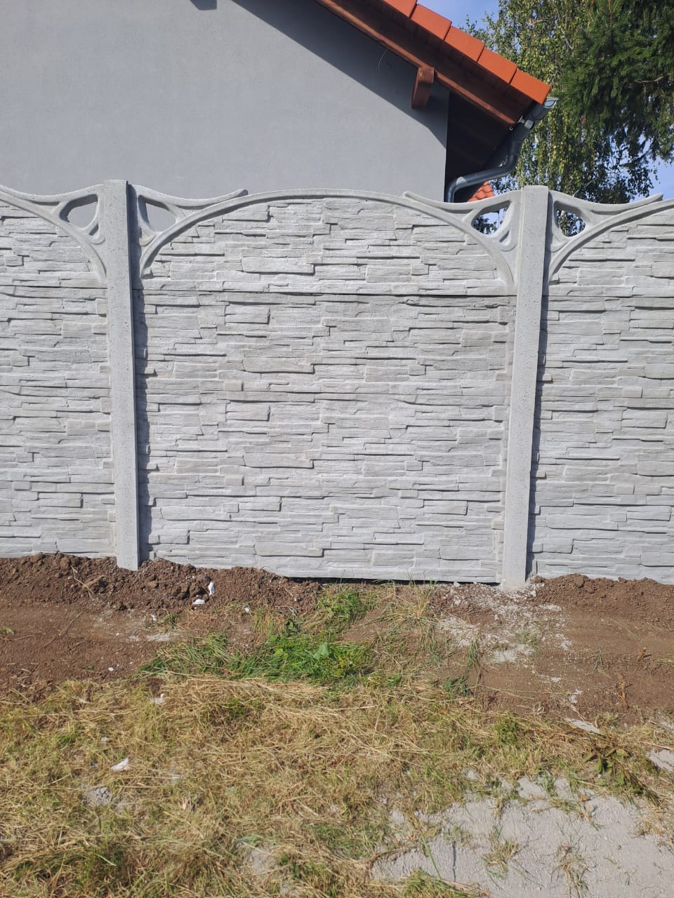 Eleganckie ogrodzenie betonowe w odcieniach szarości z dekoracyjnymi słupkami, otoczone świeżo obsypaną ziemią i trawą, z fragmentem dachu budynku w tle.