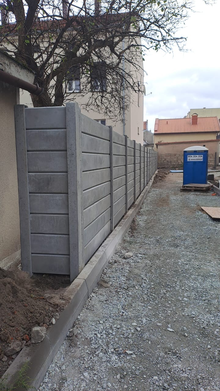 Szary, prefabrykowany płot betonowy z poziomymi panelami, widziany z boku, ciągnący się wzdłuż posesji. W tle budynki i drzewo.