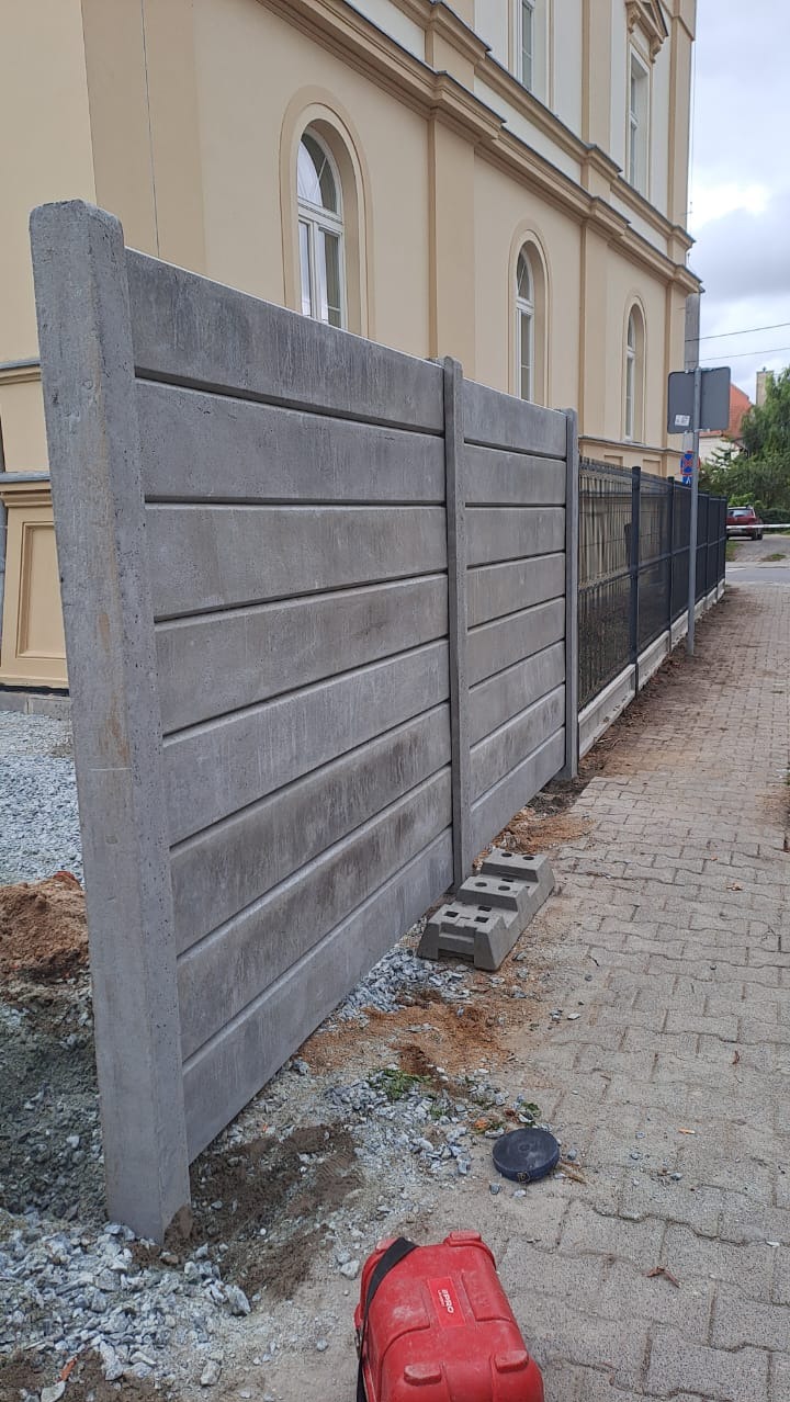 Fragment betonowego ogrodzenia panelowego z widoczną fakturą poziomych desek, z elementami montażowymi i narzędziem w tle, na tle budynku.