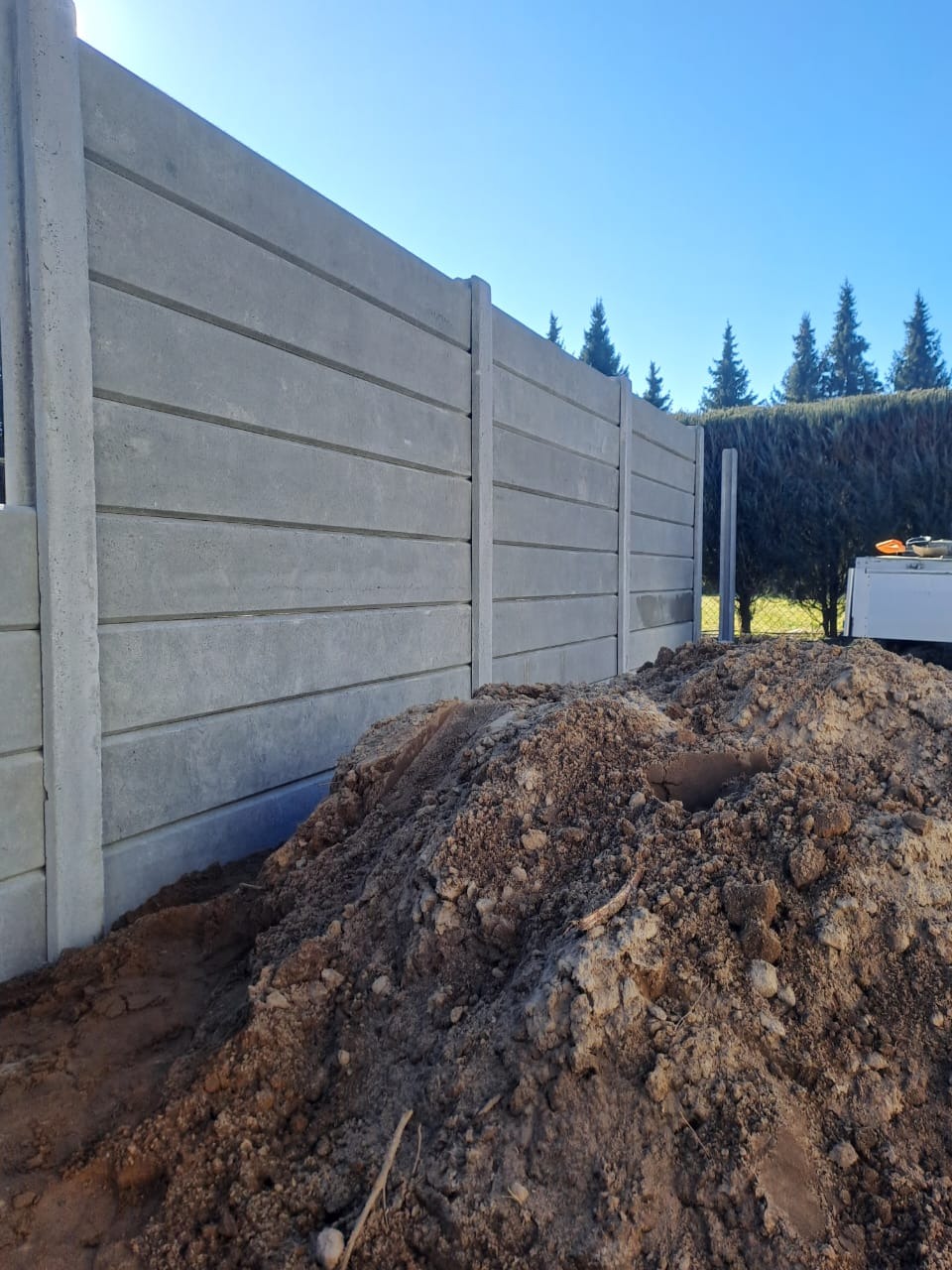 Montaż ogrodzenia betonowego w Żmigrodzie. Widoczne panele i słupki, świeżo usypana ziemia. Ujęcie z dołu, błękitne niebo w tle.