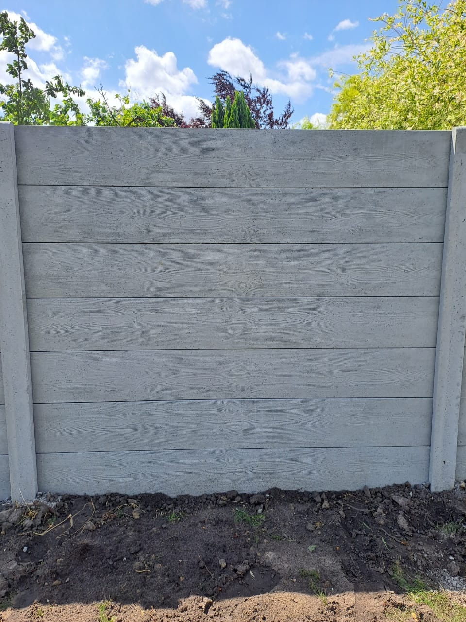 Gotowy, szary betonowy płot z imitacją drewna, z widocznymi słupkami, na tle zieleni i błękitnego nieba z chmurami.