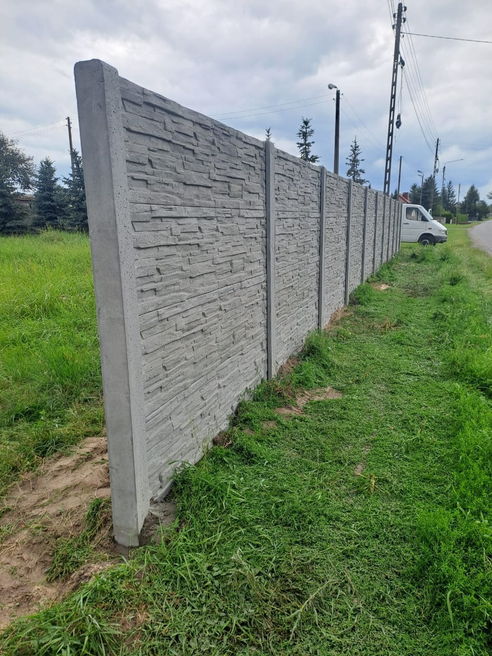 Długi, szary mur z prefabrykatów betonowych o fakturze kamienia, oddzielający posesję od drogi. Widok z poziomu gruntu, z zieloną trawą na pierwszym planie.