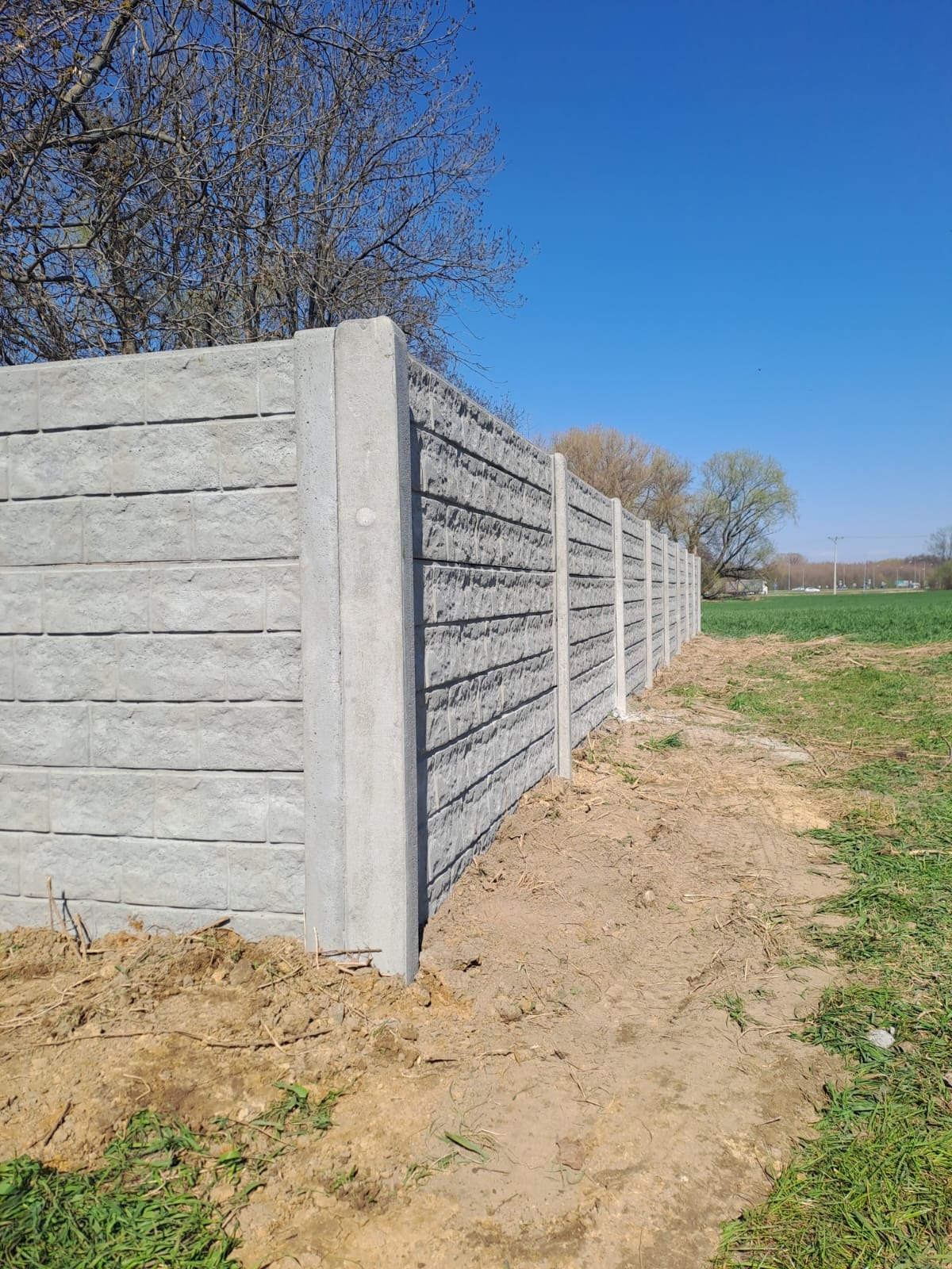 Długi, szary mur z betonowych prefabrykatów, z wyraźną fakturą, oddzielający pole od nieba. Widoczne słupy i płyty. Ujęcie z dołu, podkreślające wysokość konstrukcji.