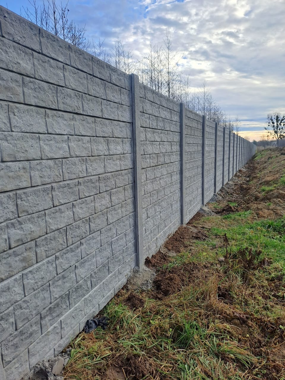 Długi, szary, betonowy płot o fakturze cegły, z widocznymi słupkami, ciągnący się wzdłuż granicy działki z trawą i ziemią, pod błękitnym niebem.